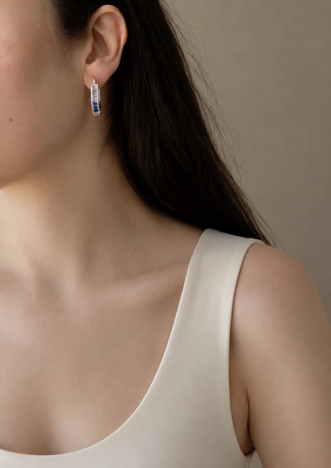 Close-up of a person wearing silver hoop earrings with blue stones against a neutral background