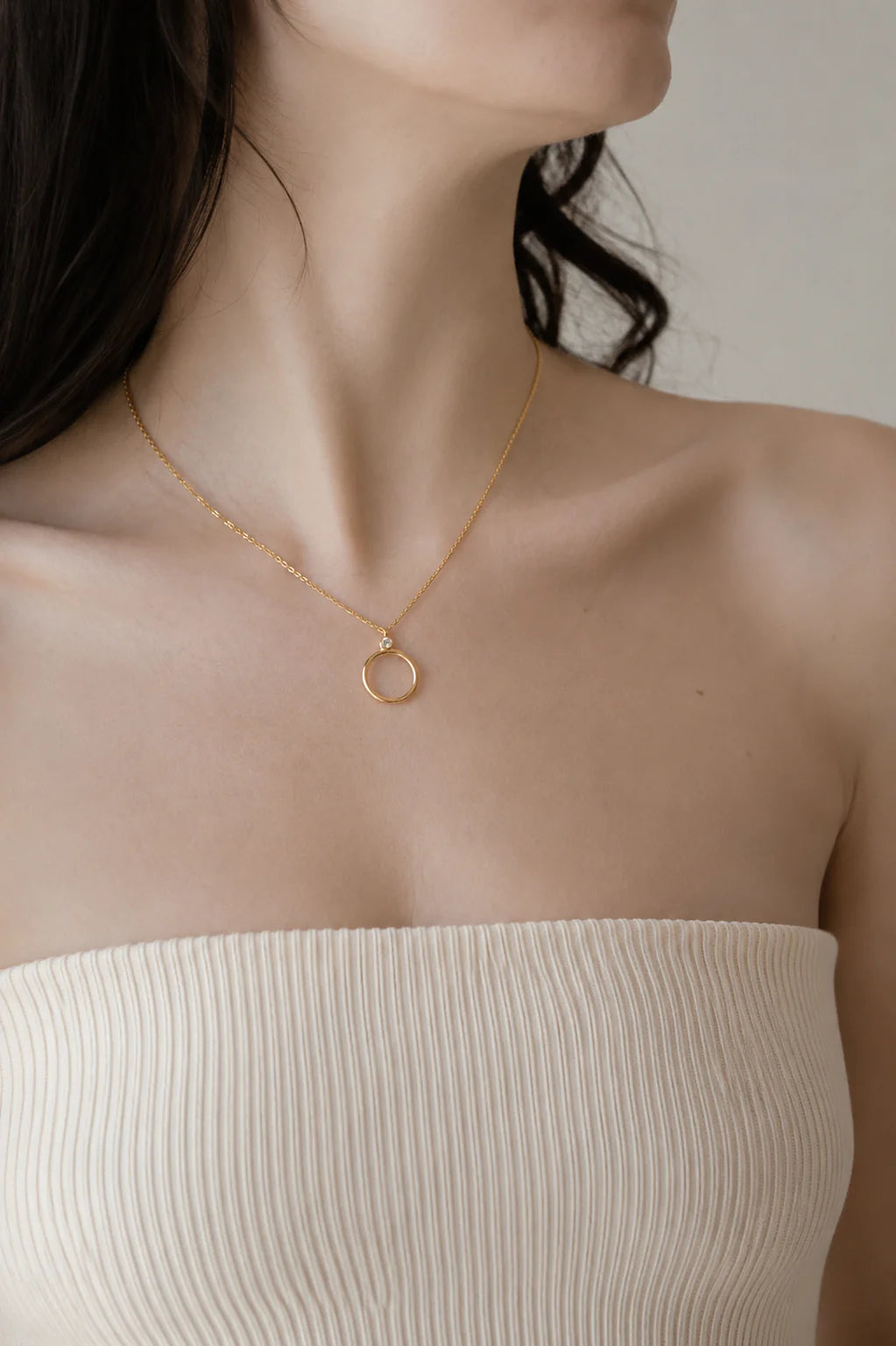 Gold necklace with a small round pendant worn by a person in a strapless beige top.